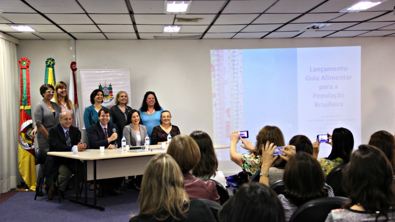 A imagem mostra os participantes do evento, de pé ou sentados atrás de uma mesa, sentados fotografados pela plateia.