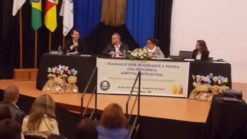 Sentado atrás da bancada, Elson segura o microfone e fala. Ao seu lado, duas mulheres e um homem. À frente, a plateia assiste sentada.