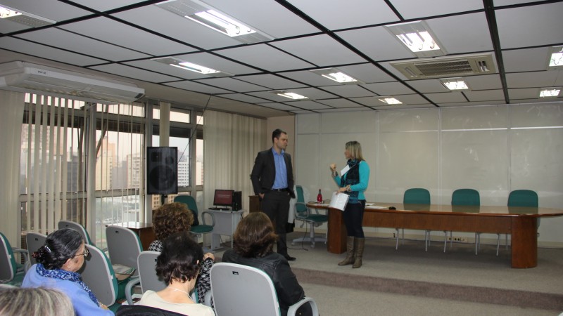 Uma mulher e um homem conversam no palco. A plateia assiste sentada.