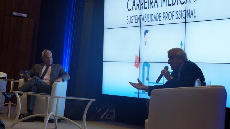 Um homem fala, segurando o microfone. O Secret&aacute;rio Gabbardo o escuta. Os dois est&atilde;o sentados em poltronas. Uma mesinha est&aacute; no meio e um tel&atilde;o est&aacute; atr&aacute;s com imagem. 