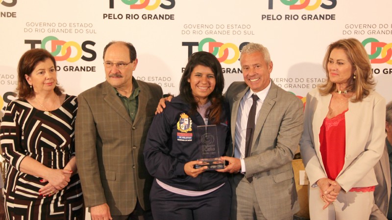 O Secretário Gabbardo abraça a vencedora e ajuda-a a segurar o troféu. O Governador Sartori e mais duas mulheres estão juntos na imagem. Todos estão de pé.