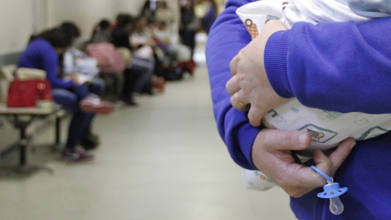 A imagem mostra uma pessoa segurando um bebê. No fundo, várias pessoas sentadas em cadeiras no corredor do hospital. 