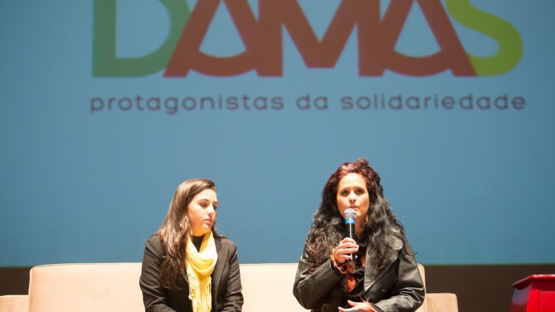 Duas mulheres sentadas no sofá, no palco. Uma fala, segurando o microfone.