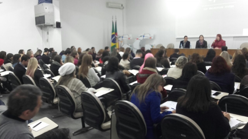 Os três palestrantes (duas mulheres e um homem) estão sentados atrás da bancada. A plateia assiste sentada.