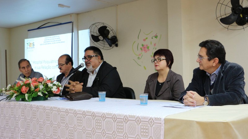 Coronel fala tendo, ao seu lado, uma mulher e três homens. Todos estão sentados atrás de uma mesa.