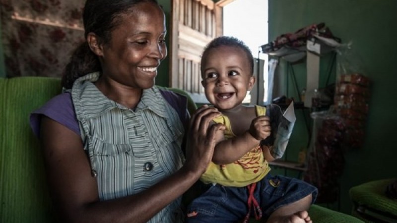 A imagem mostra uma mulher negra segurando um nenê também negro. Os dois sorriem. 