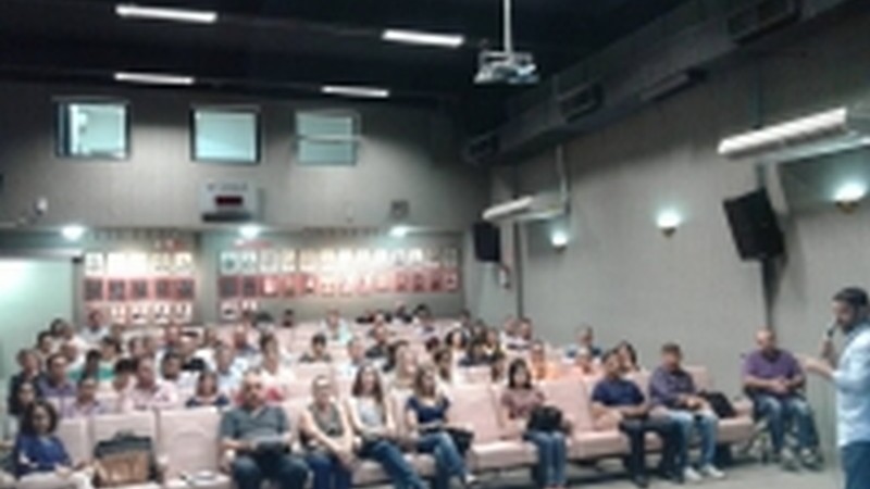 A imagem mostra o palestrante de pé, falando e segurando o microfone. No fundo, a plateia assiste sentada.