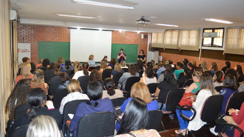 Foto tirada do fundo da sala. A palestrante fala de pé, segurando um microfone. Do seu lado, está uma mulher. A sala está lotada.  