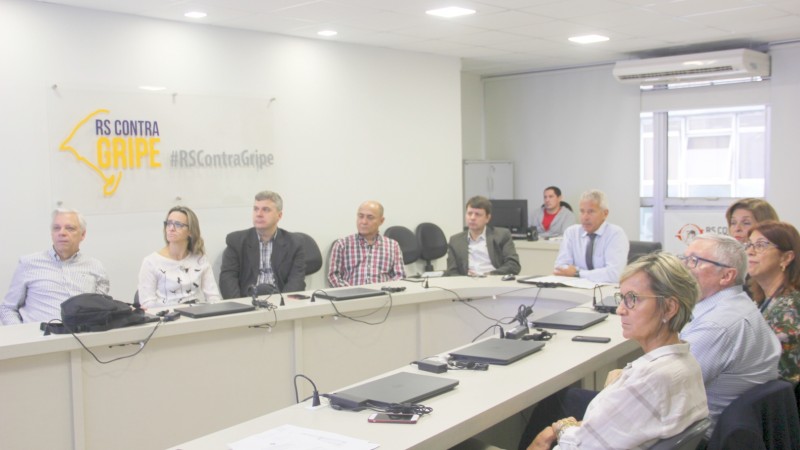 Participantes da reunião, entre eles o Secretário Gabbardo, estão sentados em torno de uma mesa, olhando para um telão.