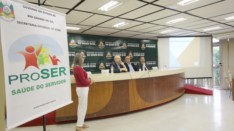 Secretário Paz fala sentado atrás da bancada, segurando o microfone. À sua esquerda, mais dois participantes do evento.