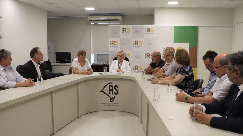 Várias pessoas sentadas, entre elas a Secretária Arita e a Diretora Ana Costa, em volta de uma mesa em U.