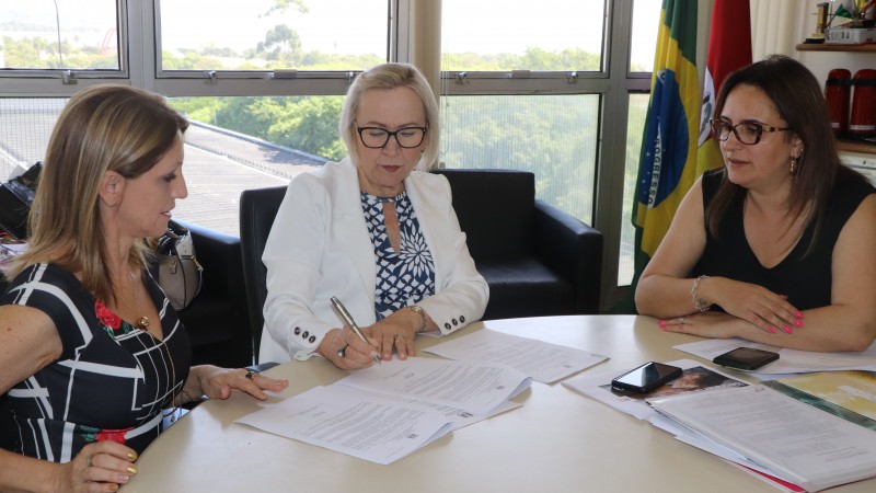 Tr&ecirc;s mulheres sentadas a uma mesa. No centro, Secretaria Arita assina um documento enquanto as outras duas observam.