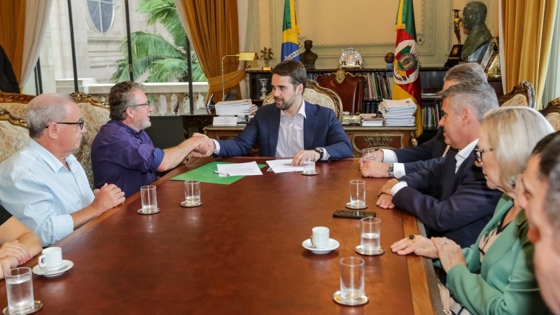 Sentados, Governador Eduardo Leite e o prefeito de São Leopoldo, Ary Vanazzi, trocam aperto de mão. A Secretária Arita e outras pessoas observam.