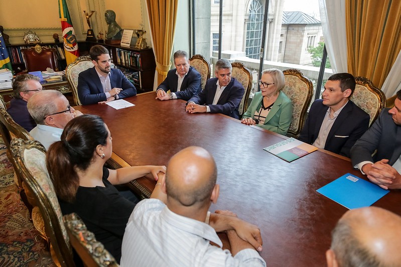 Várias pessoas sentadas em volta de uma mesa, entre elas, o Governador Leite e a Secretária Arita.