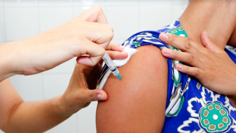 A imagem mostra as mãos de uma profissional de saúde vacinando o braço de uma mulher. 