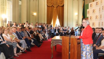 Secretária Arita de pé, falando ao microfone e atrás de um púlpito. Vê-se plateia cheia à esquerda, ao fundo da foto, e pessoas sentadas à direita.
