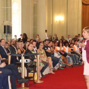 Ana Costa, de perfil, em pé, falando para um público sentado.
