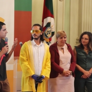 Bruno Paz fala segurando um microfone. Outras três pessoas, entre elas a Diretora Ana Costa, uma mulher e um homem ouvem. 
