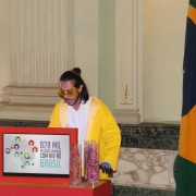 Pessoa vestindo amarelo falando. Uma tela na frente dele mostra "828 mil pessoas vivendo com HIV no Brasil".