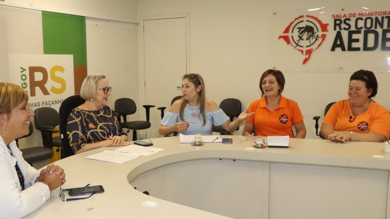 A Secretária Arita, a Diretora Ana Costa e mais três pessoas conversam, sentadas em volta de uma mesa em formato de U.