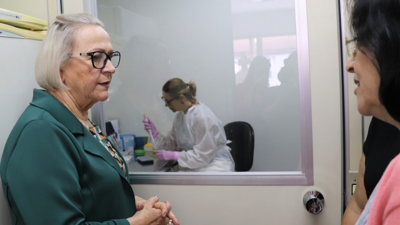 Secretária Arita conversa com uma mulher. No fundo, atrás de uma porta de vidro, uma profissional de saúde manipula amostras.  