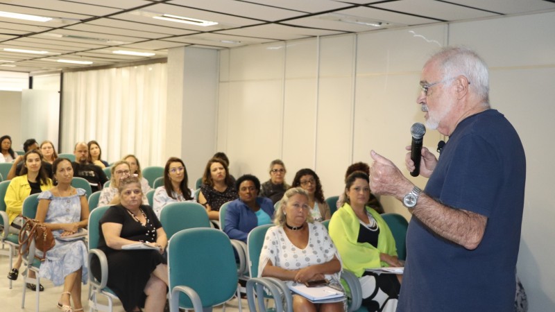 De pé, um homem fala ao microfone, atrás aparece a plateia, sentada.