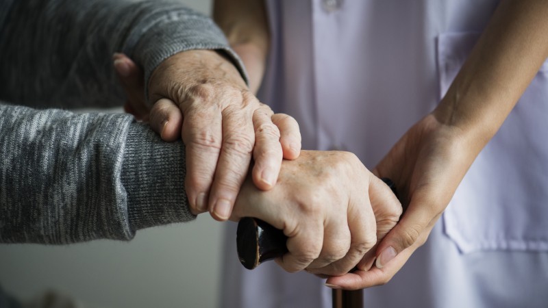 A imagem mostra duas mãos idosas segurando uma bengala com a ajuda de uma mão de uma profissional de saúde. 