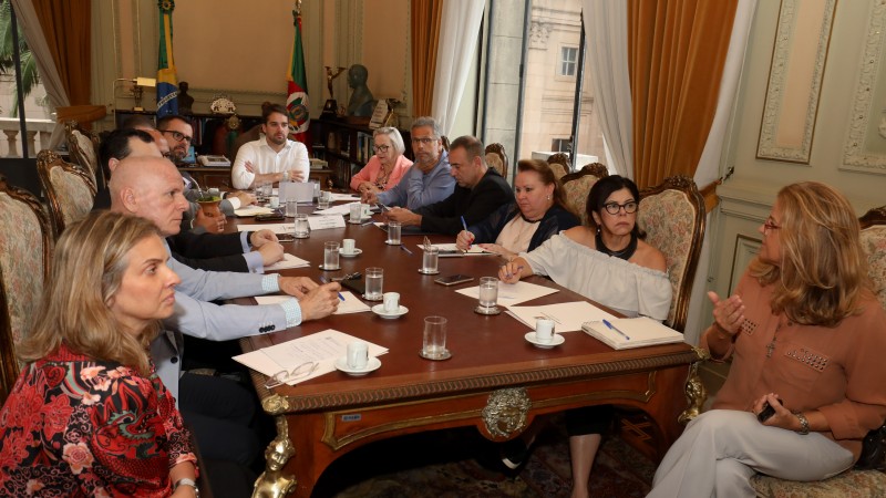 Governador Leite, Secretária Arita e vários participantes conversam em volta de uma mesa. 