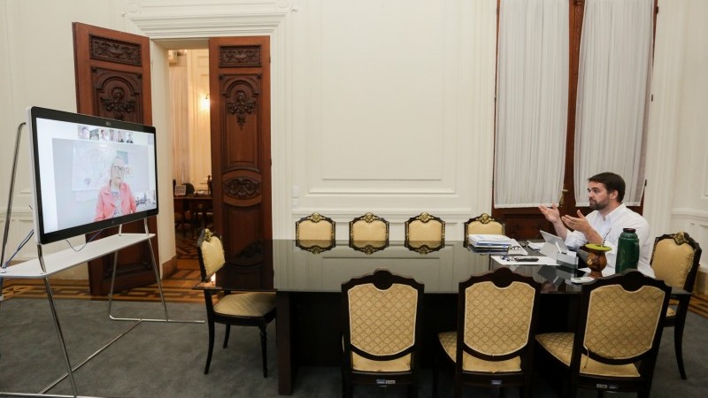 Sentado atrás de uma mesa, Governador Leite fala em videoconferência com prefeitos e Secretária Arita. 