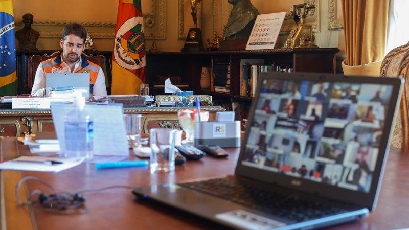 Governador Leite fala olhando para o notebook que está em cima de sua mesa. Em outra mesa, outro notebook com imagem de uma videoconferência, controles de TV, papéis e canetas.   