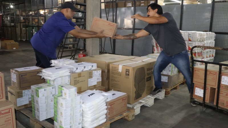Um trabalhador passa uma caixa para outro. Os dois estão de pé num depósito com outras várias caixas. 