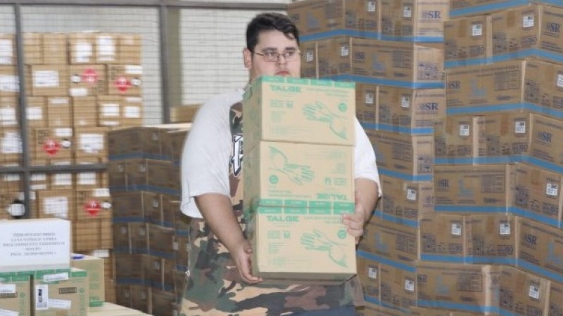 Um funcionário carrega 3 Caixas de EPI. A cena acontece num depósito com outras várias caixas. 
