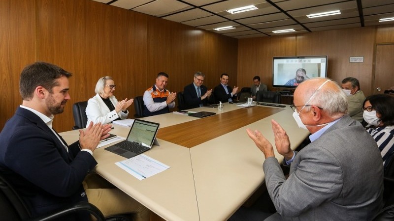 A imagem mostra o governador, secretária Arita e várias pessoas sentadas em volta de uma mesa, batendo palmas.