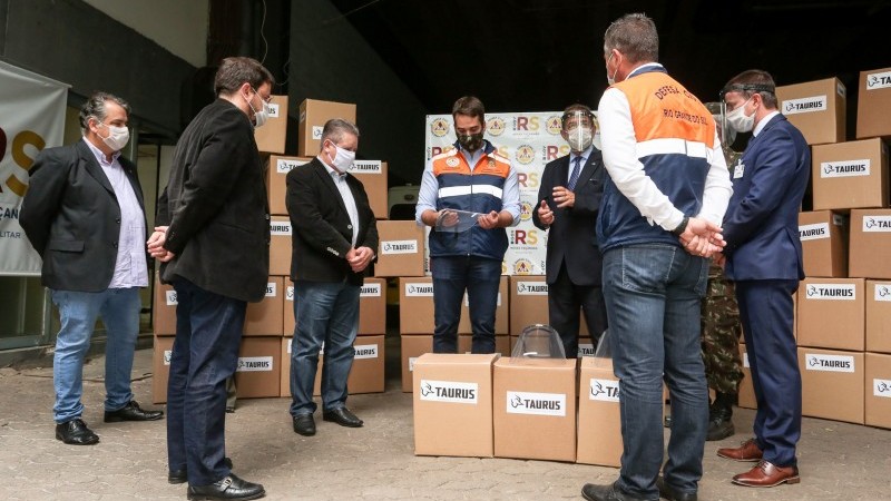 Governador Leite pega e olha um face shield enquanto sete homens o observam. Todos estão de pé e de máscara. Um deles está uniformizado de militar.