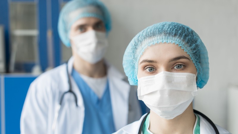 Dois profissionais de saúde (uma mulher e um homem) com máscaras, jaleco e touca.