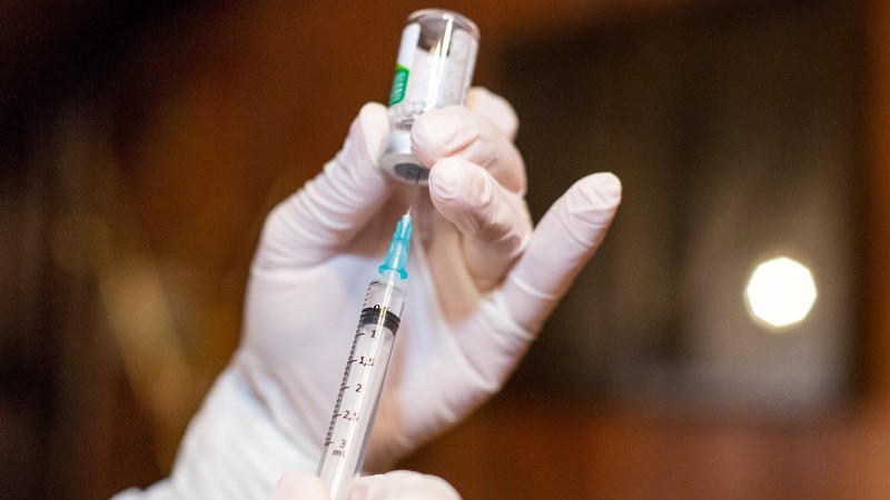 A imagem mostra duas mãos com luva preparando uma vacina. 