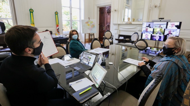 Governador, Secretária Arita e secretária-adjunta Aglaé sentados a uma mesa, com uma TV ao fundo, mostrando outras pessoas em videoconferência. Todos usam máscaras.