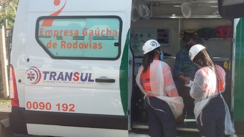 De fora da van, duas profissionais uniformizadas conversam com o caminhoneiro, que está sentado do lado de dentro do veículo.