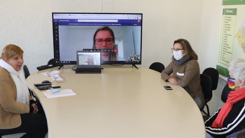 A imagem mostra três participantes da reunião conversando sentadas atrás de uma mesa e, ao fundo, um telão com a imagem da outra participante da reunião. 