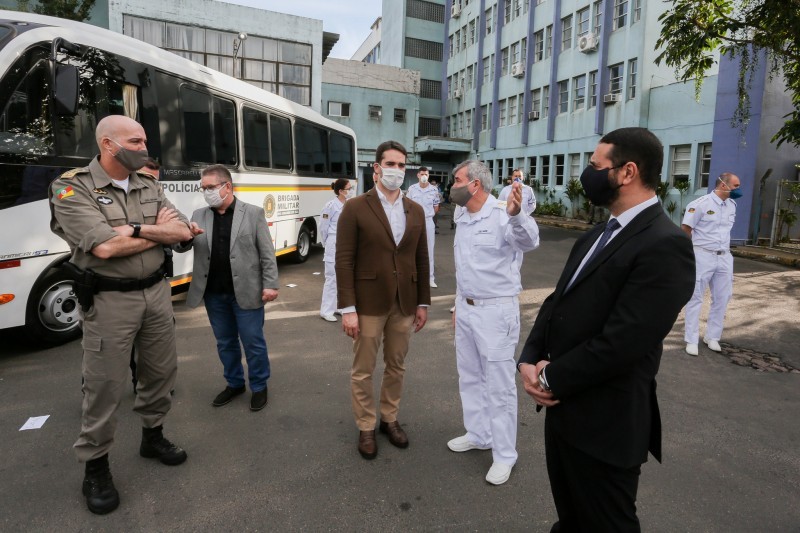 Fora do hospital, Leite recebe explicação de médico. Outros homens observam a cena. Todos estão de máscara. No fundo, um ônibus da Brigada Militar.