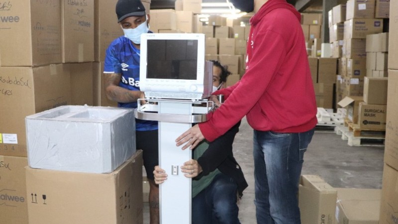 Três funcionários do setor de suprimentos organizam um equipamento no depósito, que tem várias caixas empilhadas.  