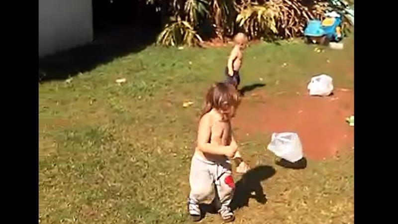 A imagem mostra duas crianças pequenas, sem camisa, brincando na grama. 