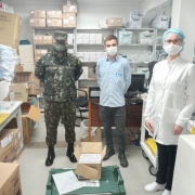 Um militar, um homem e uma profissional de saúde estão dentro de um depósito cheio de caixas. Os três estão de máscara. 