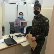 A imagem mostra uma funcionária segurando uma caixa e um militar segurando um envelope. Os dois estão de máscara e separados por um vidro. 