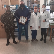 Um militar e mais três pessoas (um homem e duas profissionais de saúde) estão num depósito. Todos estão de máscara e o homem segura uma caixa com remédios. 