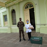 Um militar e uma profissional de saúde posam em frente ao hospital. A profissional segura uma caixa. Os dois estão de máscara. 