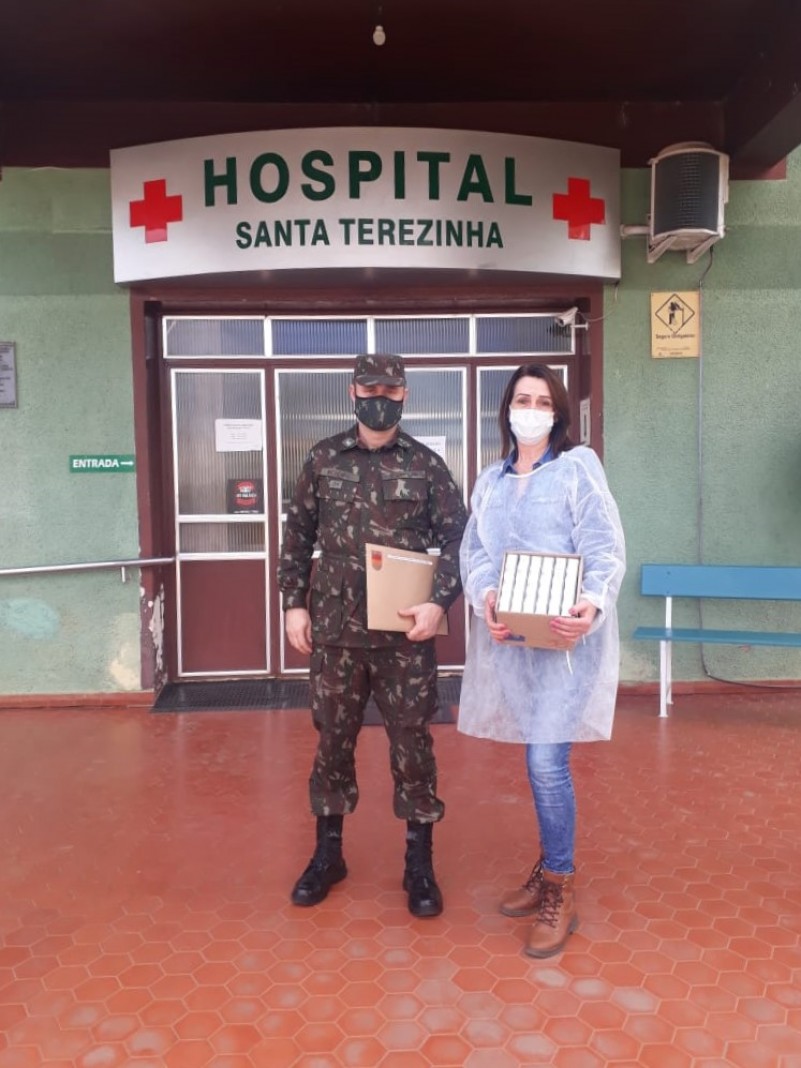 Um militar e uma profissional de saúde estão na frente da entrada do hospital. Ele segura um envelope e ela segura uma caixa com medicamentos. Os dois estão de máscara.
