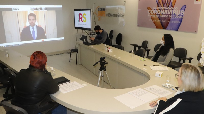 Secretária Arita e mais três participantes da reunião acompanham a fala do Governador Leite, que está projetada no telão que está a frente.