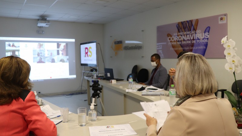 Secretária Arita e outros participantes da reunião prestam atenção na live que está sendo projetada no telão que está a frente. 