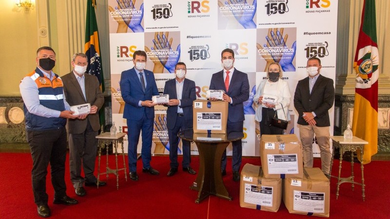 A imagem mostra o governador Leite, secretária Arita e mais cinco pessoas segurando pacotes de máscaras. Na frente deles, algumas caixas com pacotes de máscaras.